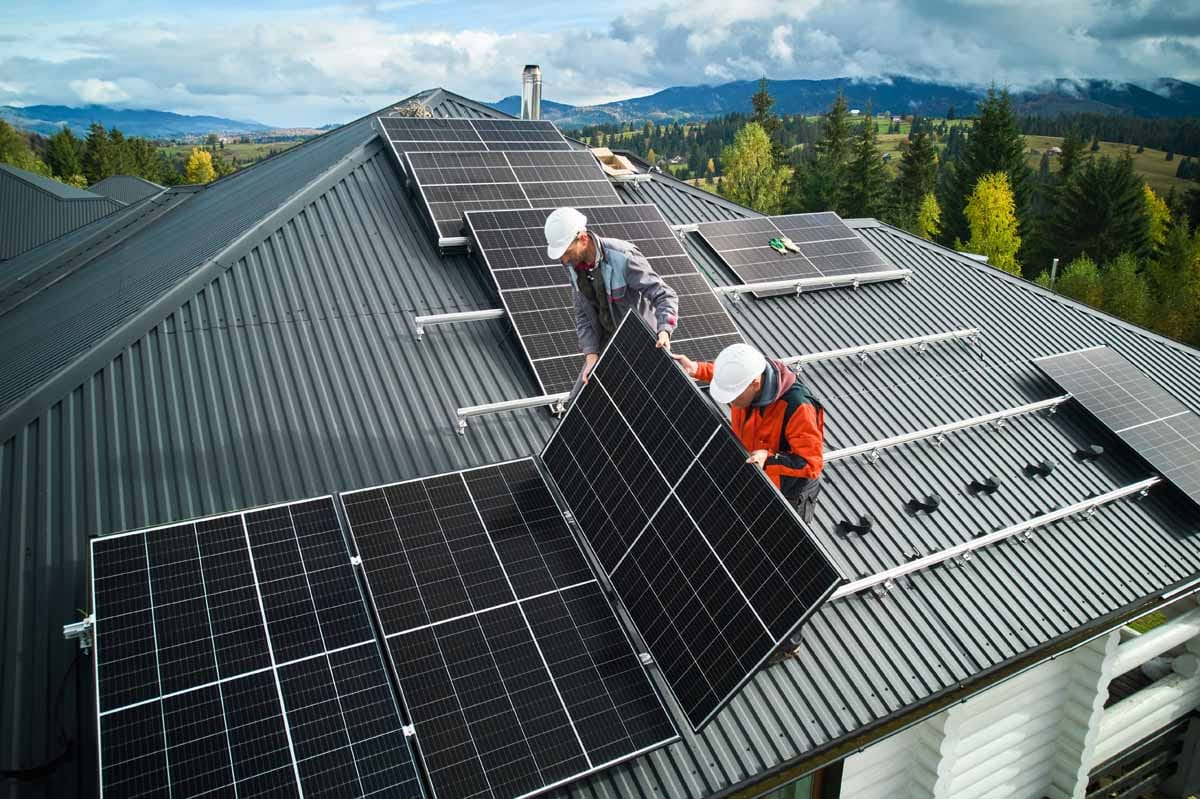Montaža sončne elektrarne na kovinski strehi – dva monterja nameščata fotovoltaični panel na nosilne tirnice; v ozadju gozd in hribi